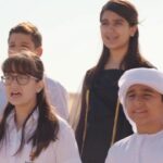 Malayali choir of 132 singers performing UAE national anthem “Eishi Biladi” in four-part harmony at Dubai Flag Garden surrounded by UAE flags.